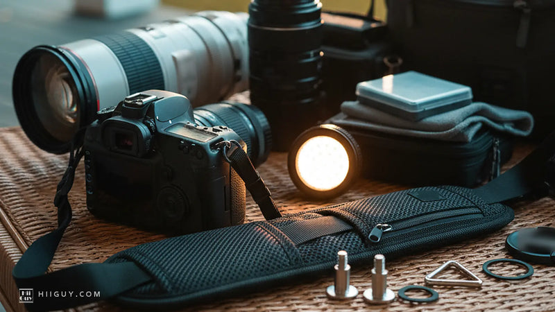 A wide padded camera strap lying face up on honey rattan at golden hour, a DSLR-style body seen from behind beside it, two steel screw studs and a split ring in front, and a small LED light burning.
