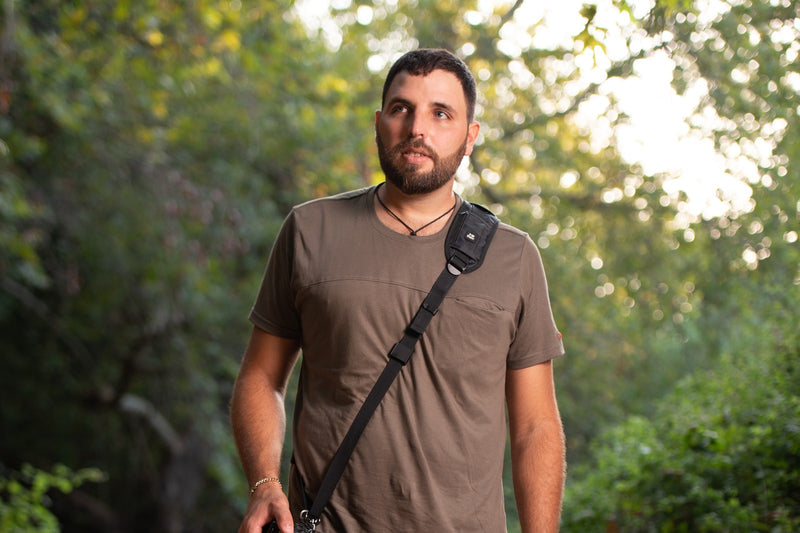 A photographer outdoors carrying a DSLR and telephoto lens on a padded HiiGuy shoulder strap