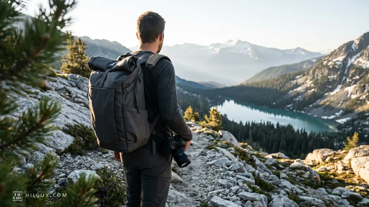 Hiker seen from behind on a rocky alpine trail in warm morning light, a dark backpack on his shoulders and a camera hanging low at his right hip under his hand, looking out over a turquoise lake and snow-dusted peaks