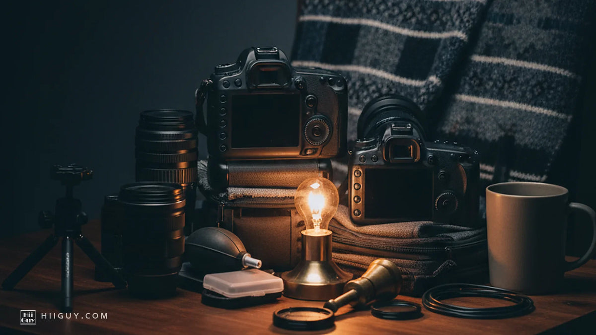 Two camera bodies of different sizes seen from behind on a dark wooden table at night, a small lamp burning between them, lenses, a blower and a mug around them, a woven blanket behind
