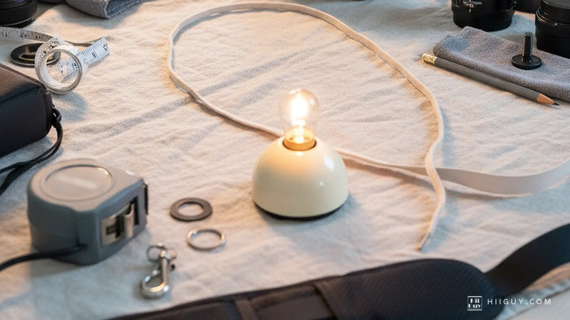 A loop of pale cord laid closed on linen around a small lit lamp, a tape measure, two split rings and a silver hook beside it, and a wide padded camera strap running across the bottom of the frame.