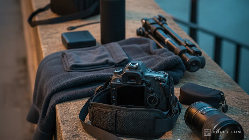 Back of a black DSLR on a stone parapet at dusk, a wide padded camera strap lying in front of it, with a fleece, a folded tripod and a lens.