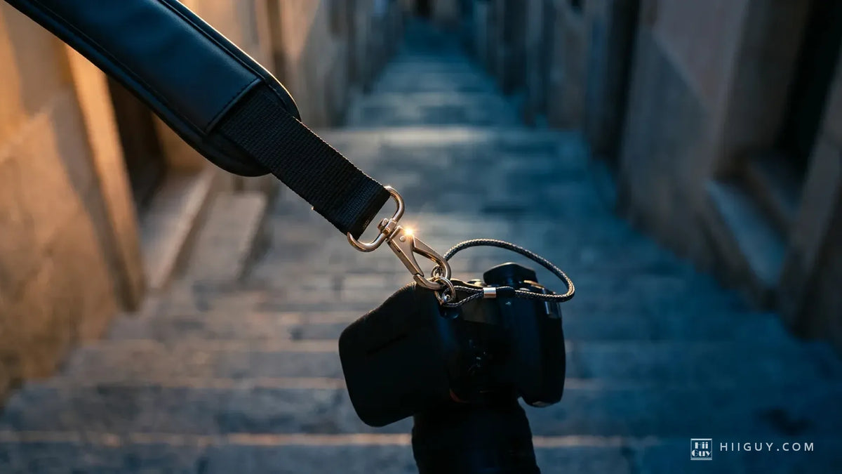 A padded black camera strap running down to a steel spring hook and a thin safety cord on a camera hanging lens-down, above a long flight of stone steps falling away out of focus in blue evening light.