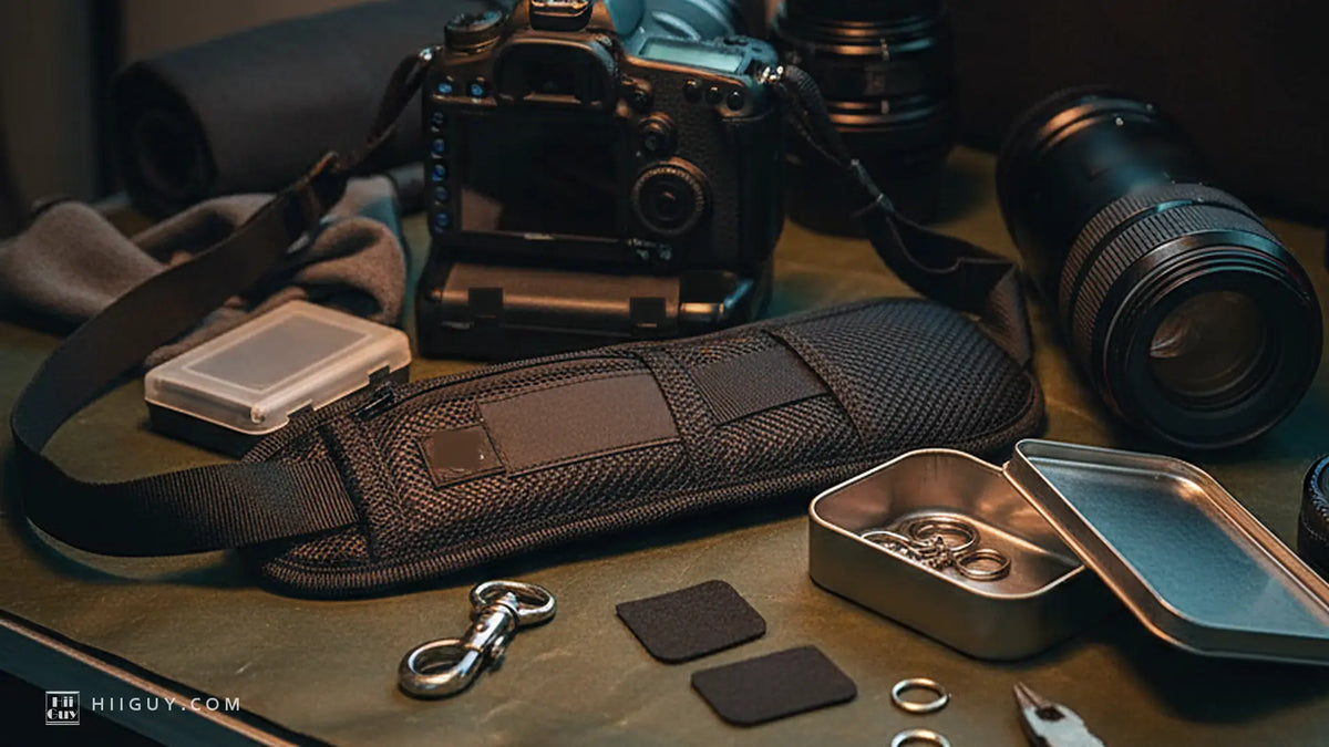 Warm lamplight on a dark bench at night: a padded black camera strap pad and its silver swivel clip, two leather tabs, split rings in an open metal tin, a DSLR and lenses behind.