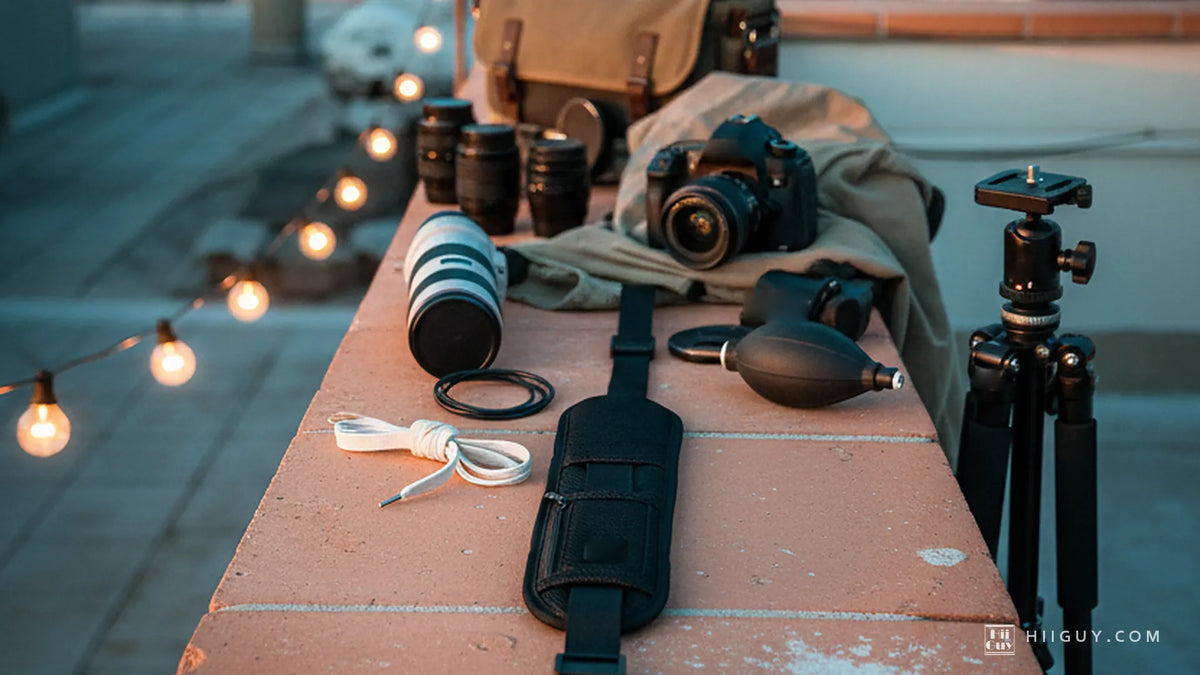 Camera strap laid out at full length on a warm terracotta rooftop ledge at dusk, lenses and a camera behind it, string lights lit.