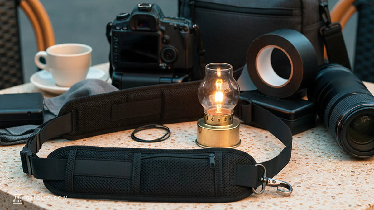 A wide padded camera strap laid in a loop on a speckled stone cafe table at dusk, a lit brass lantern beside it, a camera, bag and telephoto lens behind in the blue city light