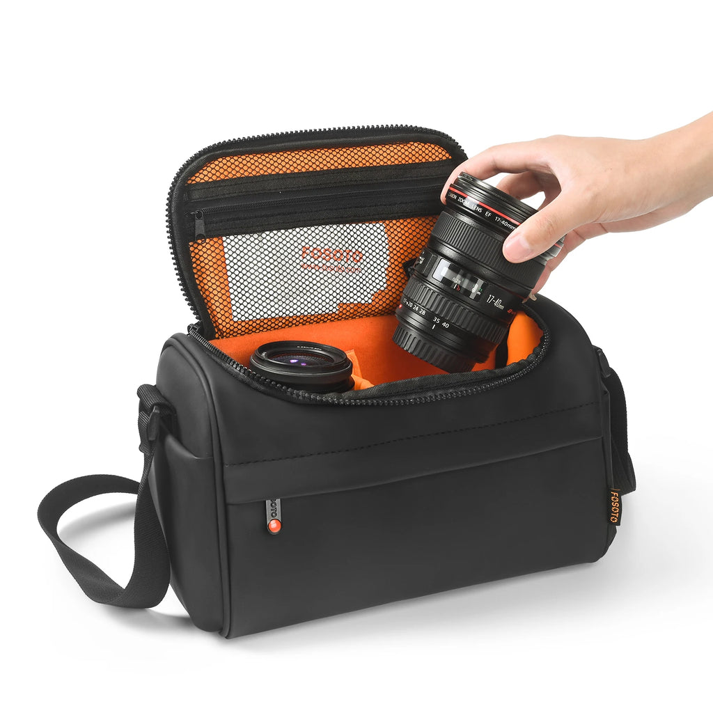 Black camera shoulder bag open to an orange padded interior, a small lens inside and a hand lowering a zoom lens in