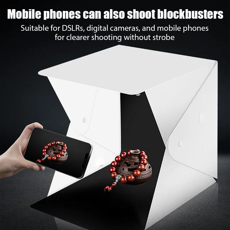 Hands holding a DSLR in front of a lit white folding light box with gold beaded bracelets on its floor