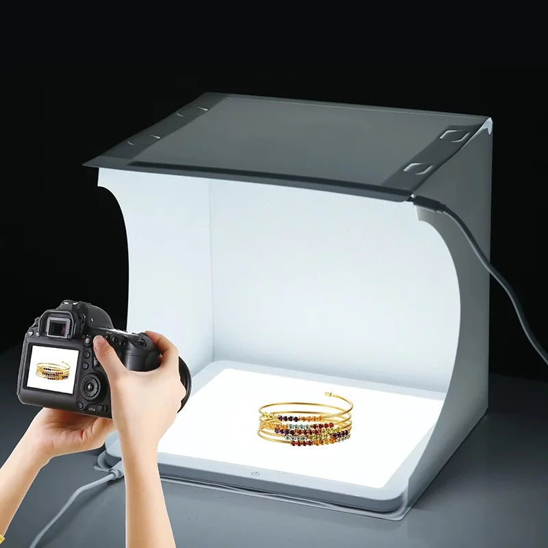 Hands holding a DSLR in front of a lit white folding light box with gold beaded bracelets on its floor
