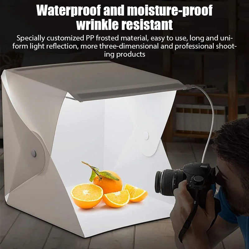 Hands holding a DSLR in front of a lit white folding light box with gold beaded bracelets on its floor