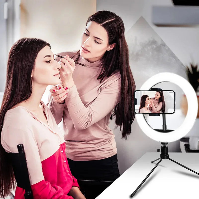 LED ring light on a small black tripod holding a phone on a makeup table, with a woman applying blusher behind it
