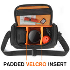 Black camera shoulder bag open to an orange padded interior, a small lens inside and a hand lowering a zoom lens in