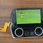 PSP Go console lying face up with an orange ribbon adapter attached at the left edge and a Memory Stick entry on screen