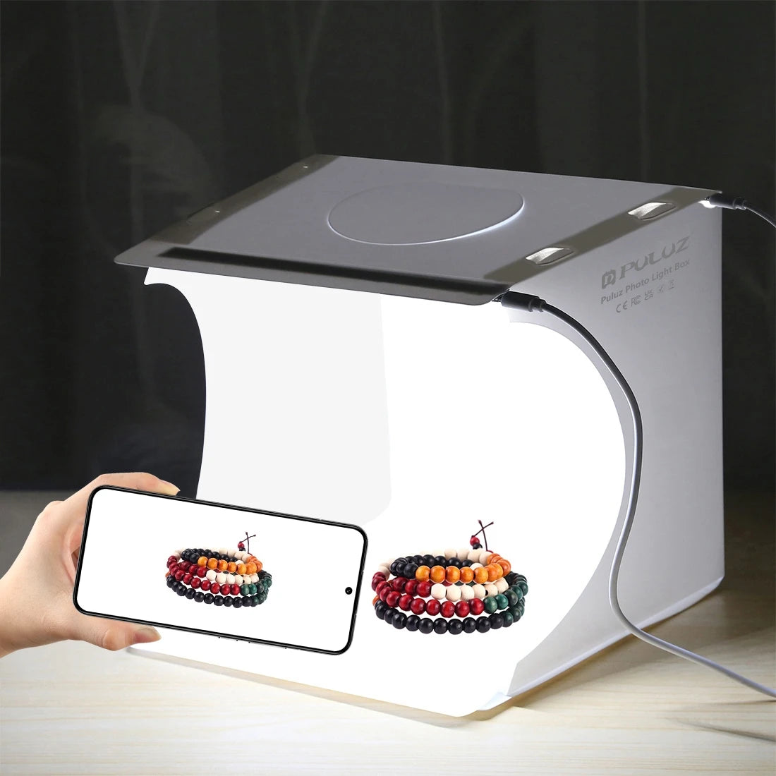Small white Puluz folding light box lit from above with beaded bracelets inside, and a hand holding a phone showing the shot