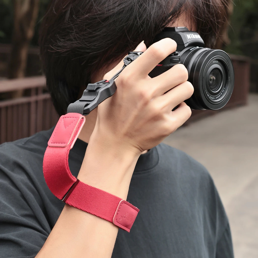 Orange camera wrist strap with a black clip worn on a wrist, the hand holding a black mirrorless camera