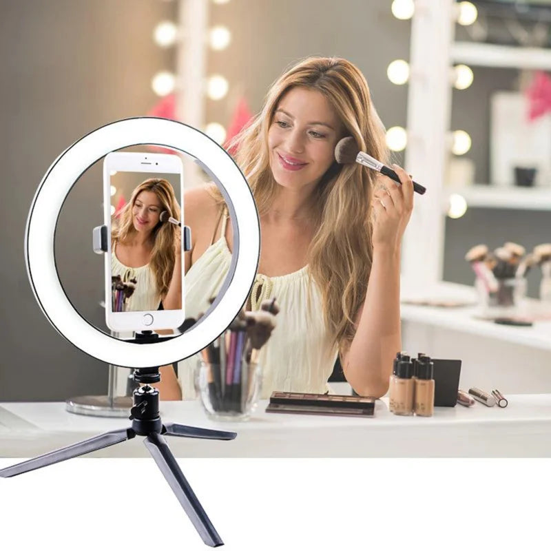 LED ring light on a small black tripod holding a phone on a makeup table, with a woman applying blusher behind it