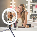 LED ring light on a small black tripod holding a phone on a makeup table, with a woman applying blusher behind it
