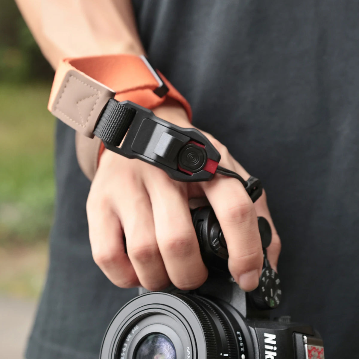 Orange camera wrist strap with a black clip worn on a wrist, the hand holding a black mirrorless camera